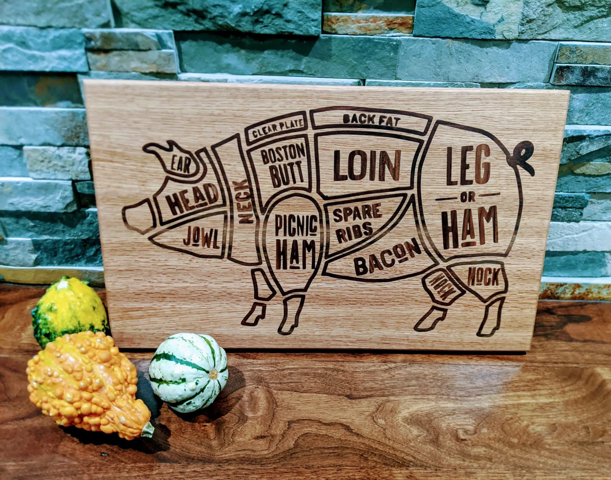 A wooden pig diagram cutting board labeled with pork cuts, such as loin, leg or ham, bacon, and ribs. Three decorative gourds are in front, and the background is a stone tile wall.