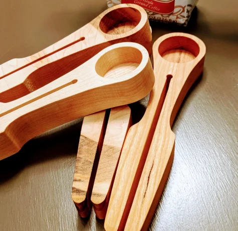 Three wooden coffee scoops with built-in bag clips are stacked on a brown surface. A package of ground coffee is partially visible in the background.
