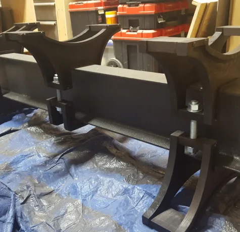 A metal and plastic assembly with bolts and curved supports sits on a blue tarp in a workshop, with toolboxes and wooden boards in the background.