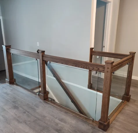 A modern staircase railing with wooden posts and handrails, featuring clear glass panels, is installed on a gray wood floor against light gray walls in a residential interior.