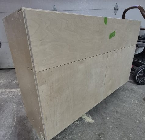 A large, unfinished wooden cabinet with clean lines sits on a concrete floor in a garage. The cabinet has two lower doors and an upper section secured with green tape. A stroller is partially visible nearby.