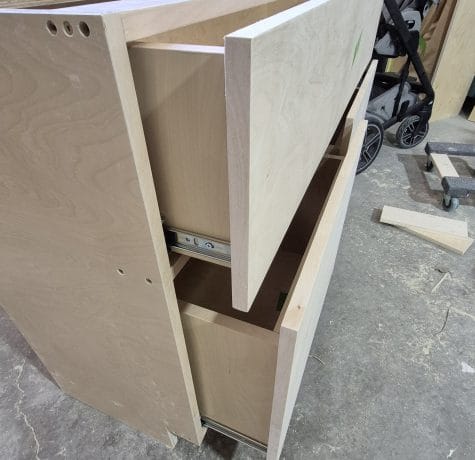 A wooden cabinet with two drawers partially open sits on a concrete garage floor. A baby stroller and some lumber are visible in the background.