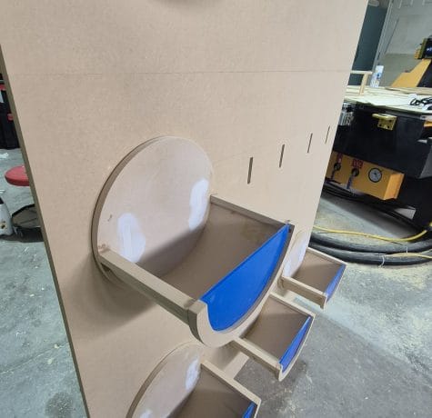 A vertical wooden board with several circular and rectangular shelves attached, some with blue painters tape. The setup is in a workshop with tools and equipment visible in the background.