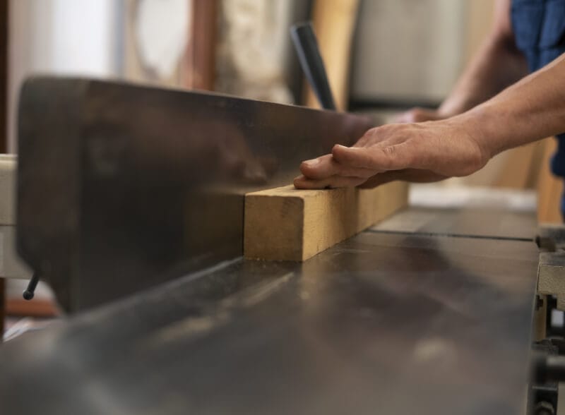 Close-up of a person’s hands guiding a wooden plank through a table saw in a workshop, demonstrating woodworking and careful handling of tools.