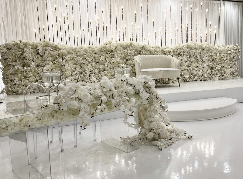 Elegant wedding stage with white floral arrangements, a cream-colored loveseat, glass tables, candles, and cascading white flowers, all set against a shimmering white backdrop.